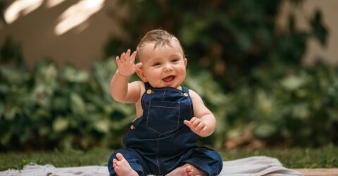 Jeune enfant assis au sol en salopette bleue, main levée, grand sourire